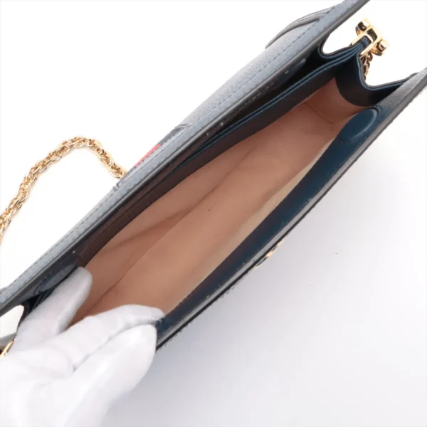 Open blue leather handbag with beige interior lining, held open by a gloved hand, showing empty compartment and a gold chain strap.