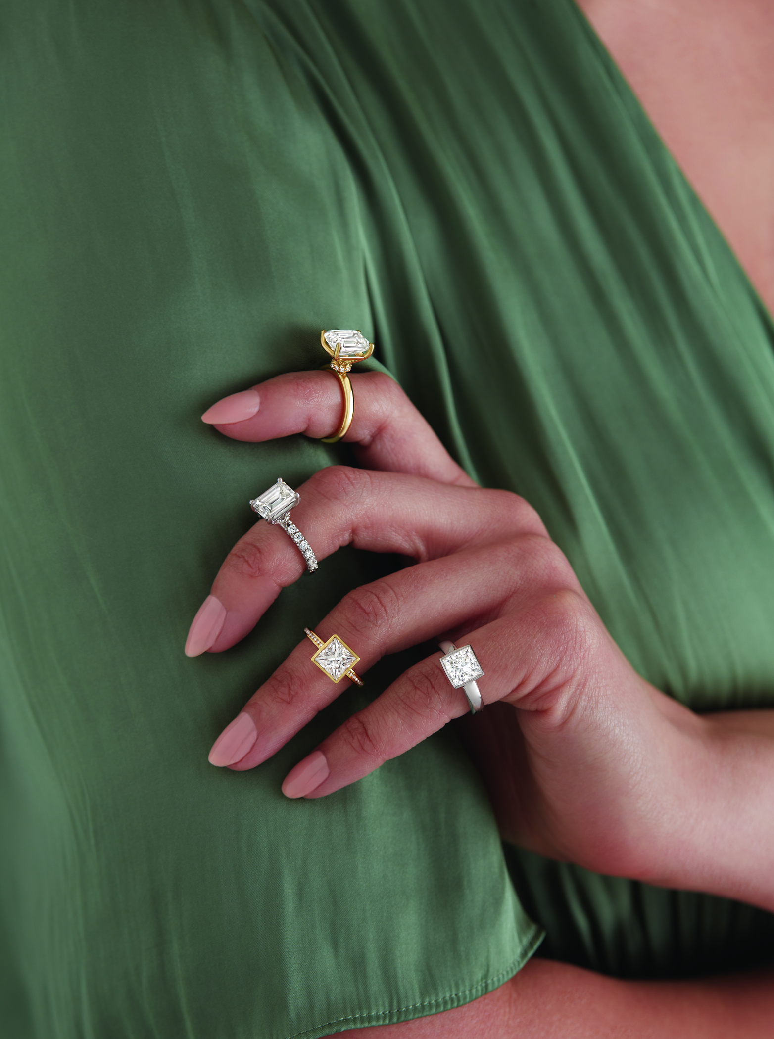 Close-up of a hand with manicured nails resting on green fabric, wearing four rings with large square and rectangular gemstones in gold and silver settings.