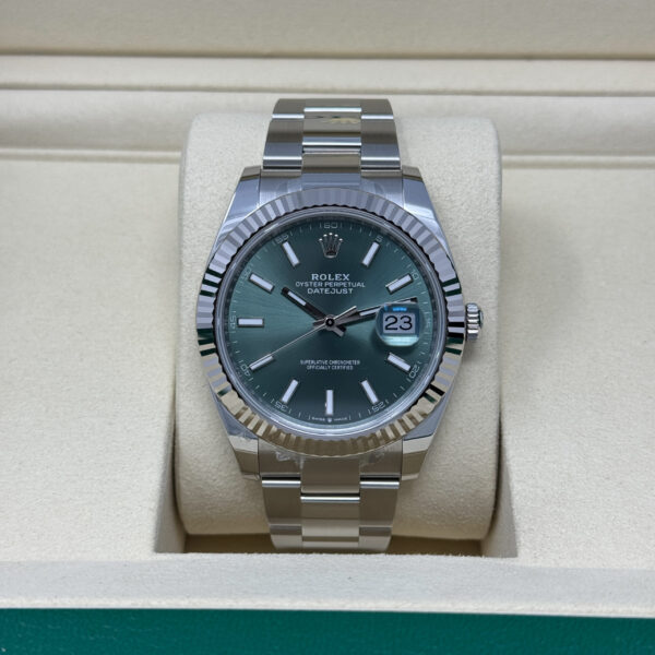 Stainless steel Rolex Datejust with fluted bezel and green sunburst dial showing date "23" on a cream watch cushion