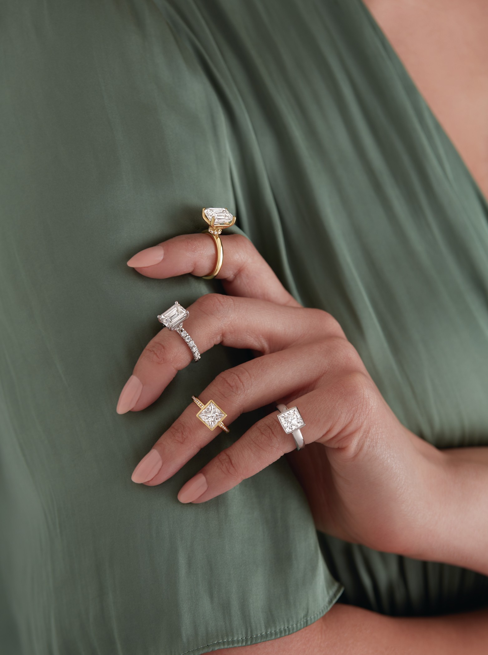 Close-up of a hand with manicured nails resting on green fabric, wearing four rings with large square and rectangular gemstones in gold and silver settings.